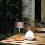 SC Hospitality Olivia Mini table lamp in corten finish, a cordless rechargeable light on a wooden outdoor table.