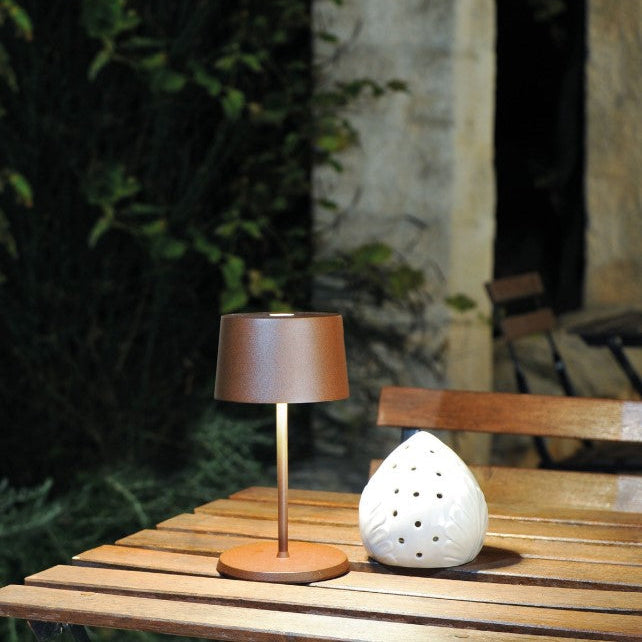 SC Hospitality Olivia Mini table lamp in corten finish, a cordless rechargeable light on a wooden outdoor table.