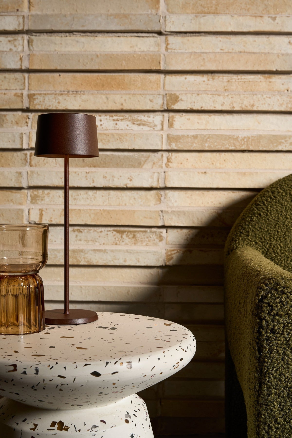 SC Hospitality Olivia Pro cordless table lamp in corten finish on a terrazzo side table next to a green armchair.