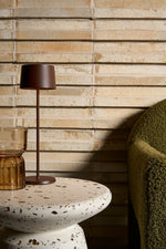 SC Hospitality Olivia Pro cordless table lamp in corten finish on a terrazzo side table next to a green armchair.