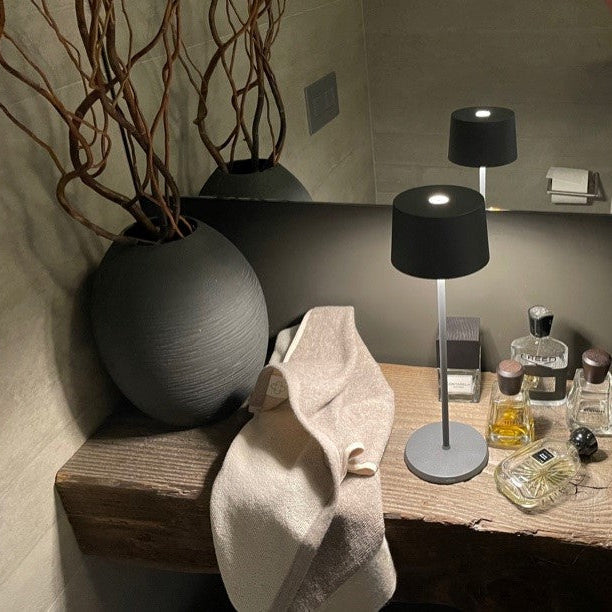 SC Hospitality Olivia Pro dark grey cordless LED table lamp on a wooden shelf next to a vase and perfume bottles