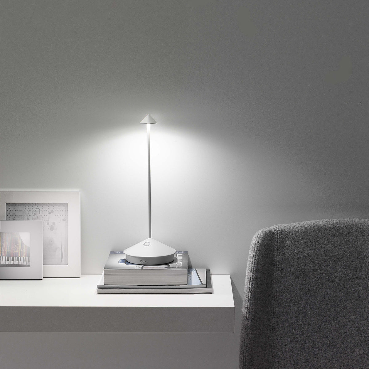 SC Hospitality Pina white cordless rechargeable table lamp glowing on a stack of books on a white shelf