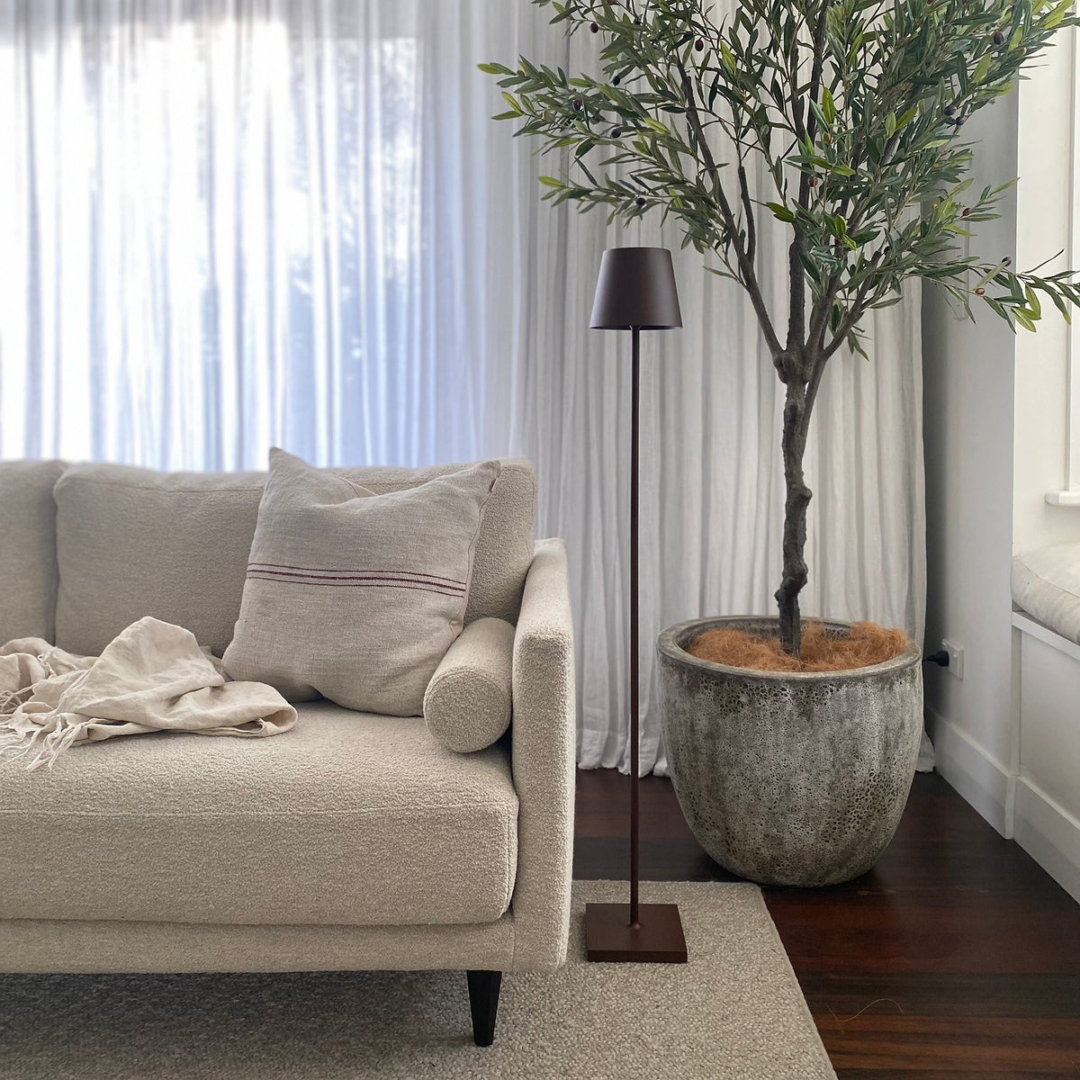 SC Hospitality Poldina Pro cordless floor lamp in corten finish standing next to a cream sofa and potted olive tree