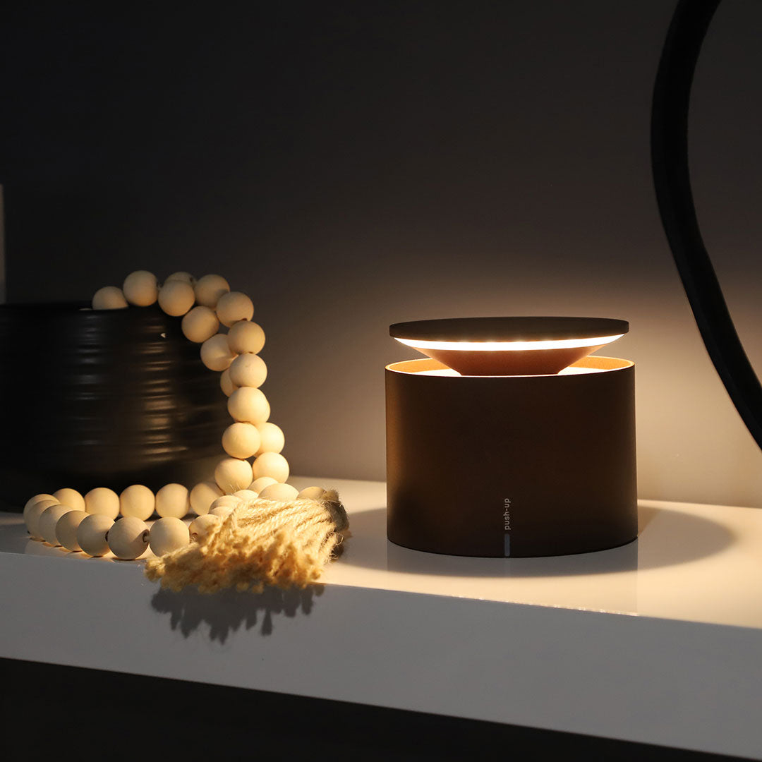 SC Hospitality Zafferano Push-Up cordless table lamp in corten finish glowing on a white shelf with wooden beads.