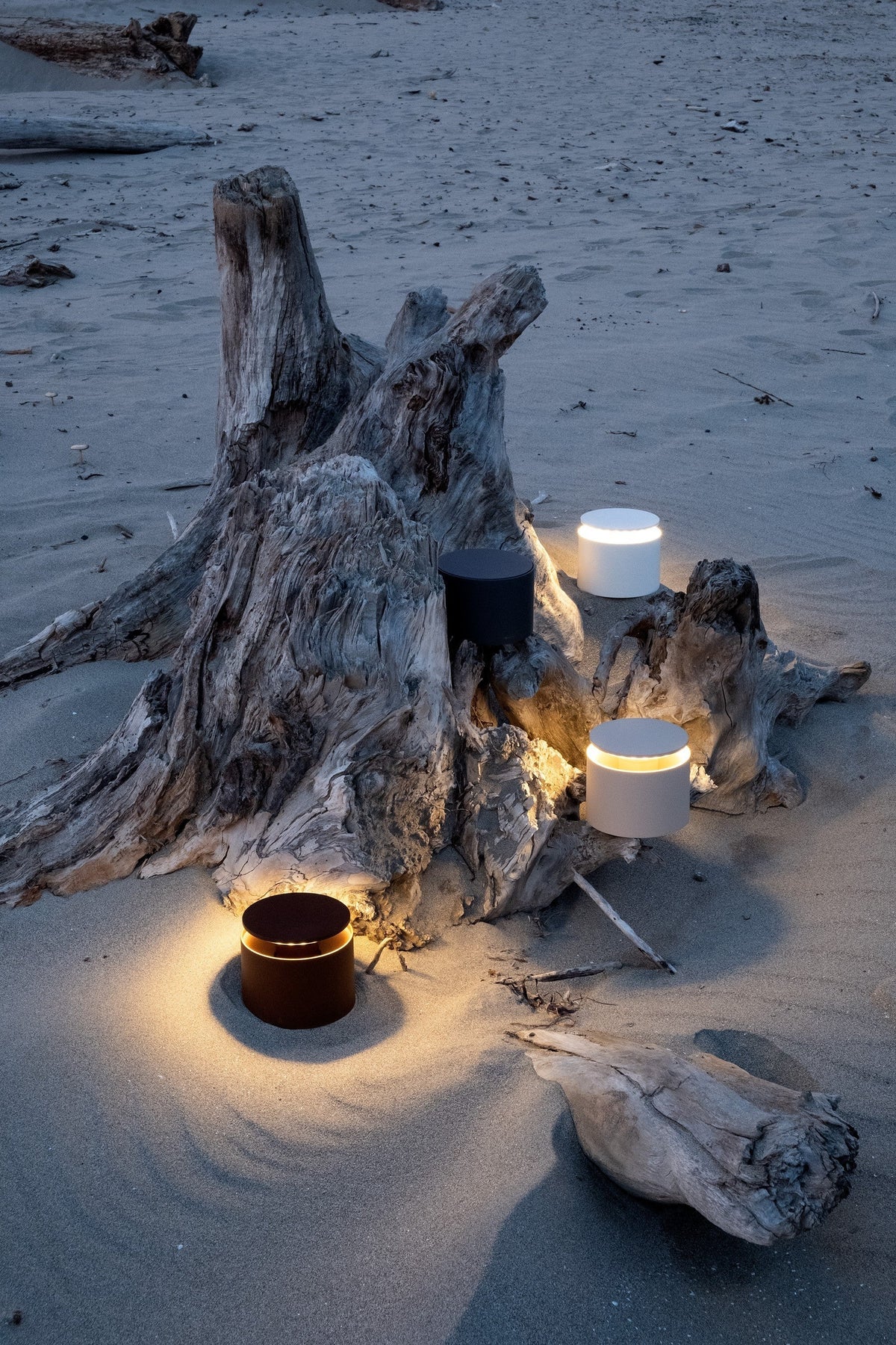 SC Hospitality Push-Up table lamps in sand, white, and black glowing on a sandy beach near driftwood at dusk.