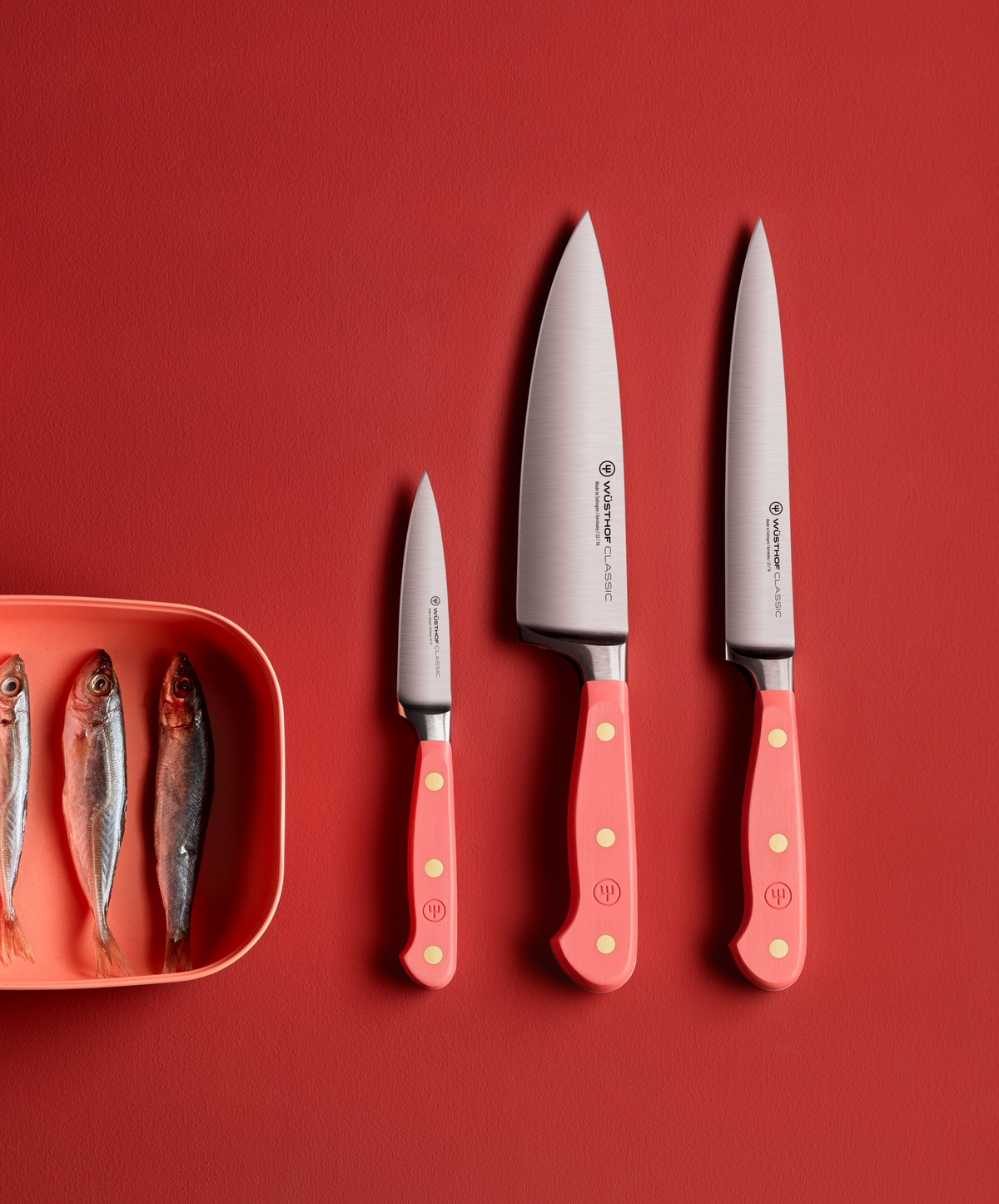 SC Hospitality Wüsthof paring and chef knives with coral peach handles next to a bowl of small fish on a red surface.