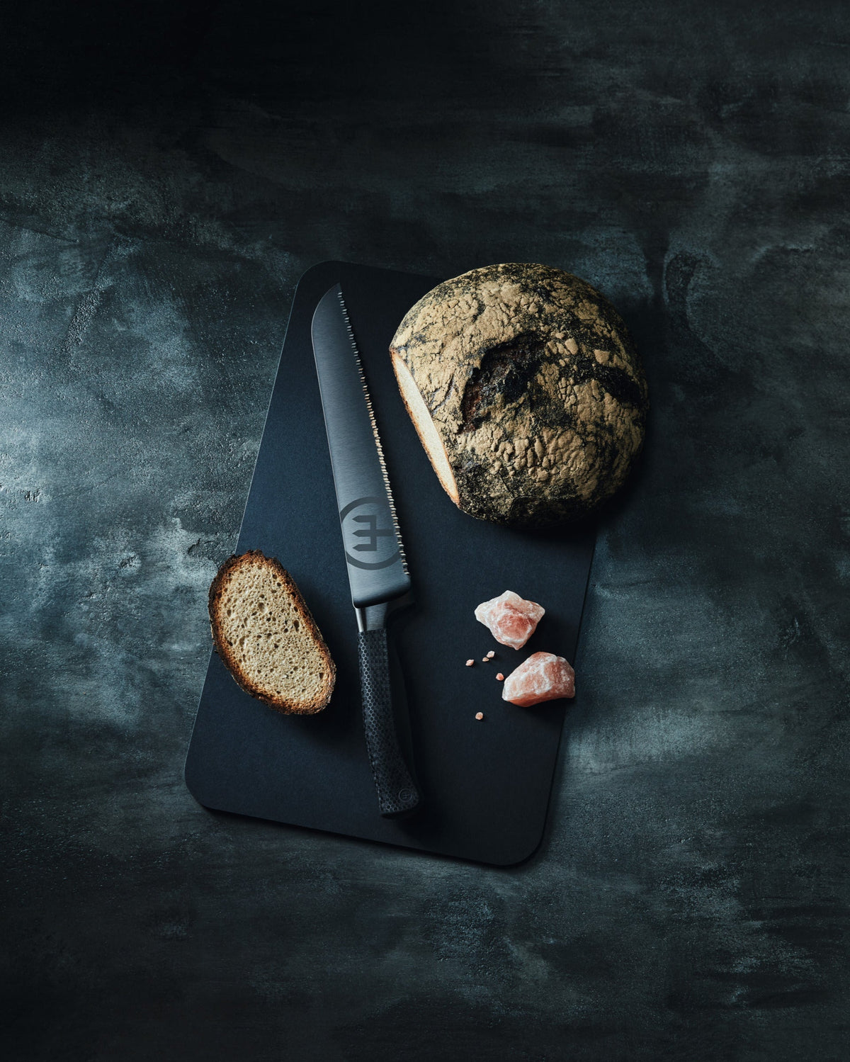 SC Hospitality WÜSTHOF Performer 23cm bread knife with black DLC coating and textured handle on a black cutting board.